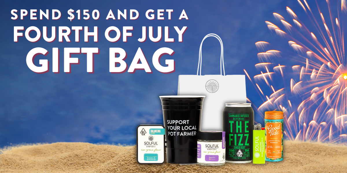 Gift bag sits on sand with assorted products in front of fireworks.